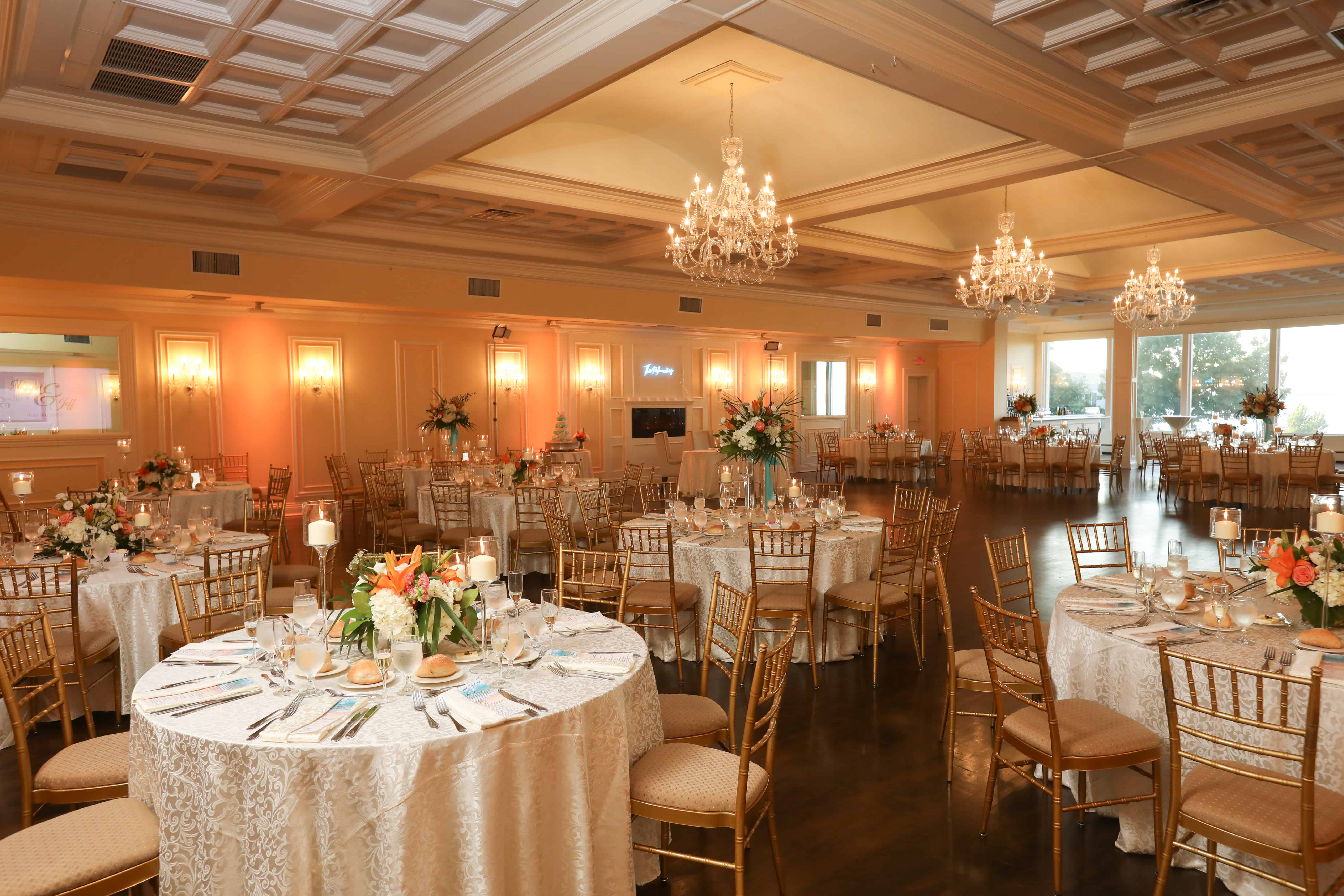 Elegant banquet hall set for an event, featuring round tables with lace tablecloths, gold Chiavari chairs, and floral centerpieces. Chandeliers hang from the ceiling, and warm lighting creates an inviting atmosphere—one of the finest wedding venues Long Island NY has to offer.