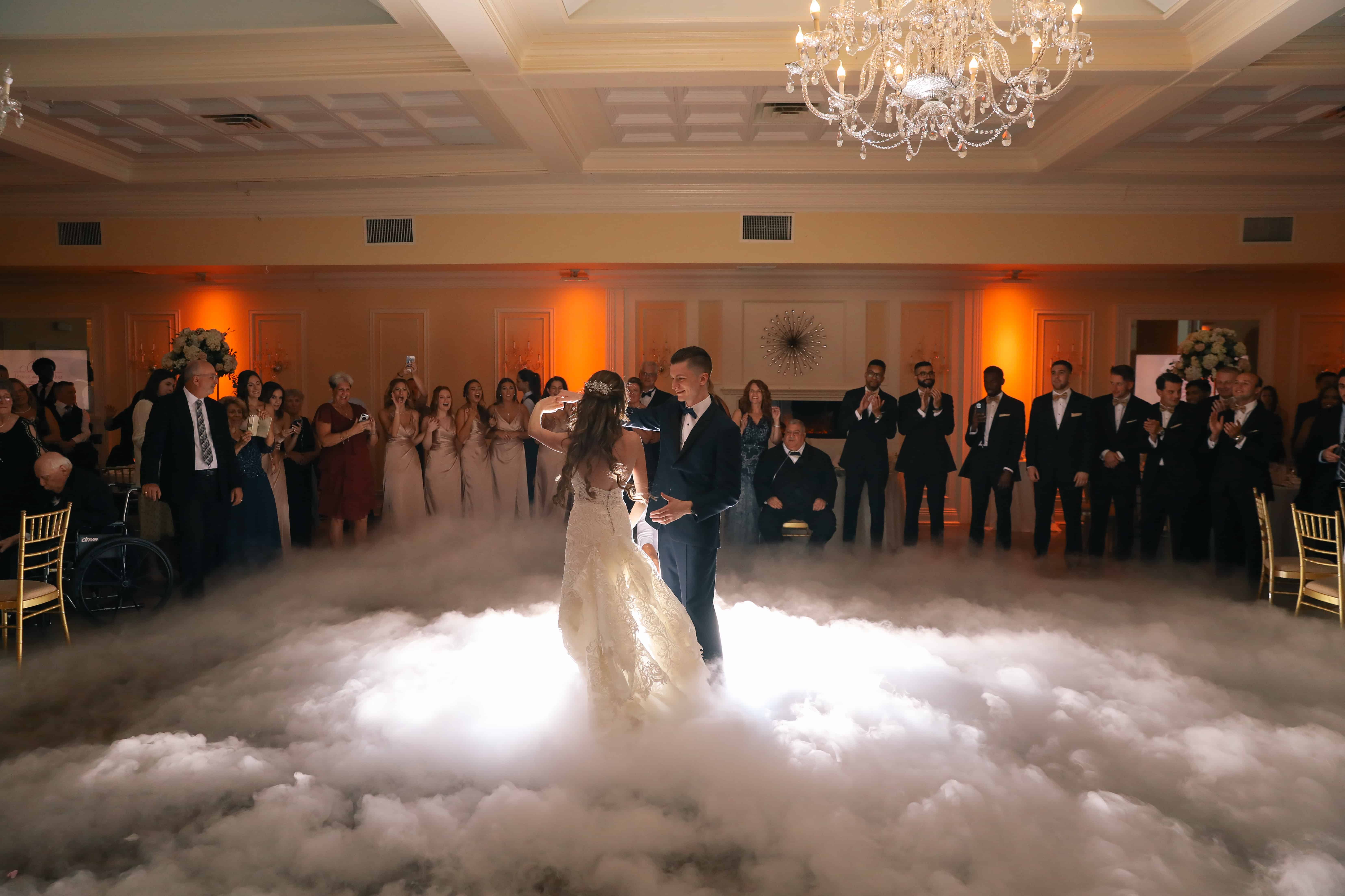 A bride and groom dance gracefully at one of the stunning wedding venues on Long Island, NY. They are enveloped in a foggy embrace, surrounded by elegantly dressed guests. A chandelier glistens above as warm orange lights cast an inviting glow upon the scene.