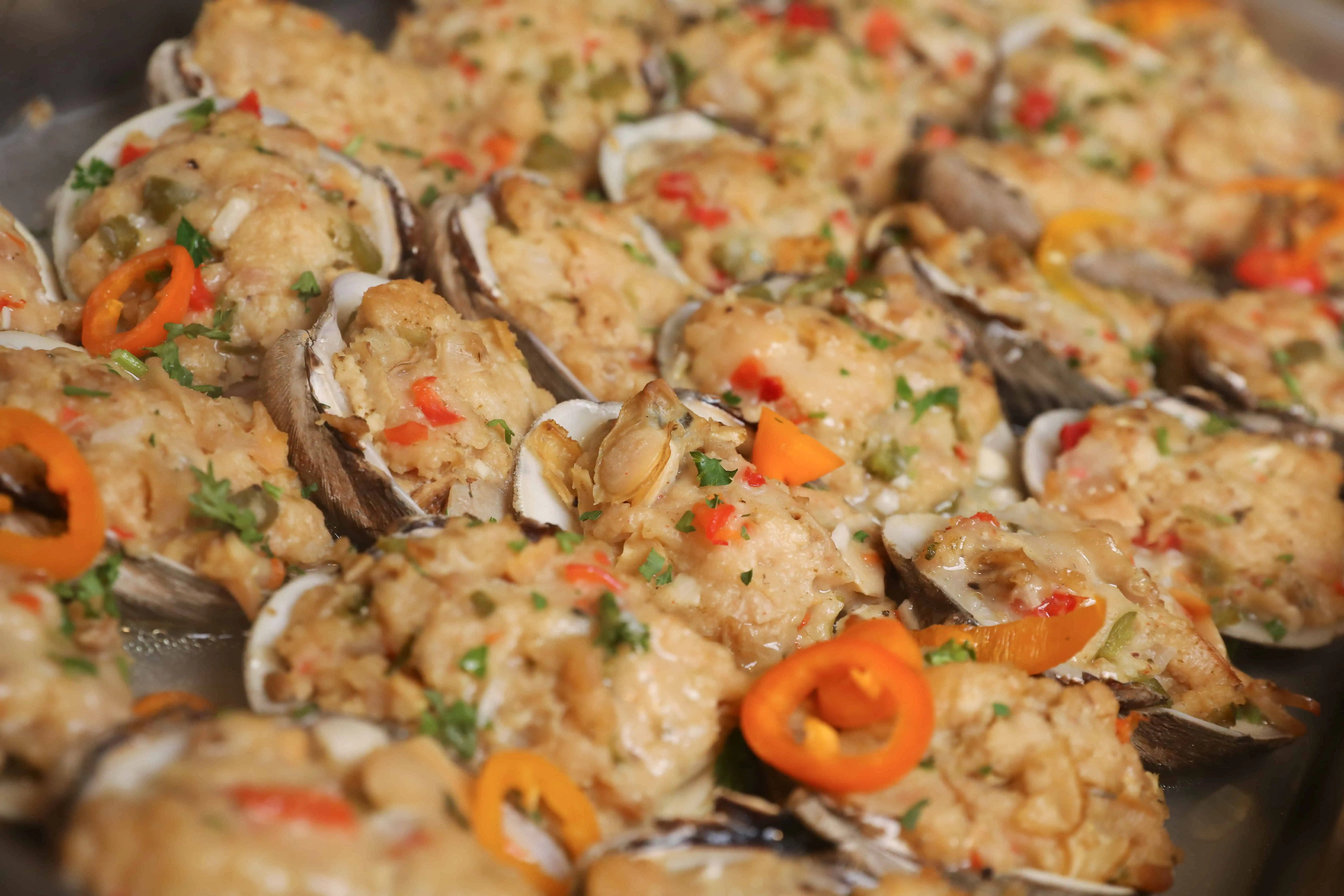 A tray of stuffed clams topped with chopped herbs and diced red and orange bell peppers graces the table at wedding venues on Long Island, NY. The golden, crumbly filling is garnished with parsley and small slices of orange pepper, creating a vibrant and appetizing presentation.