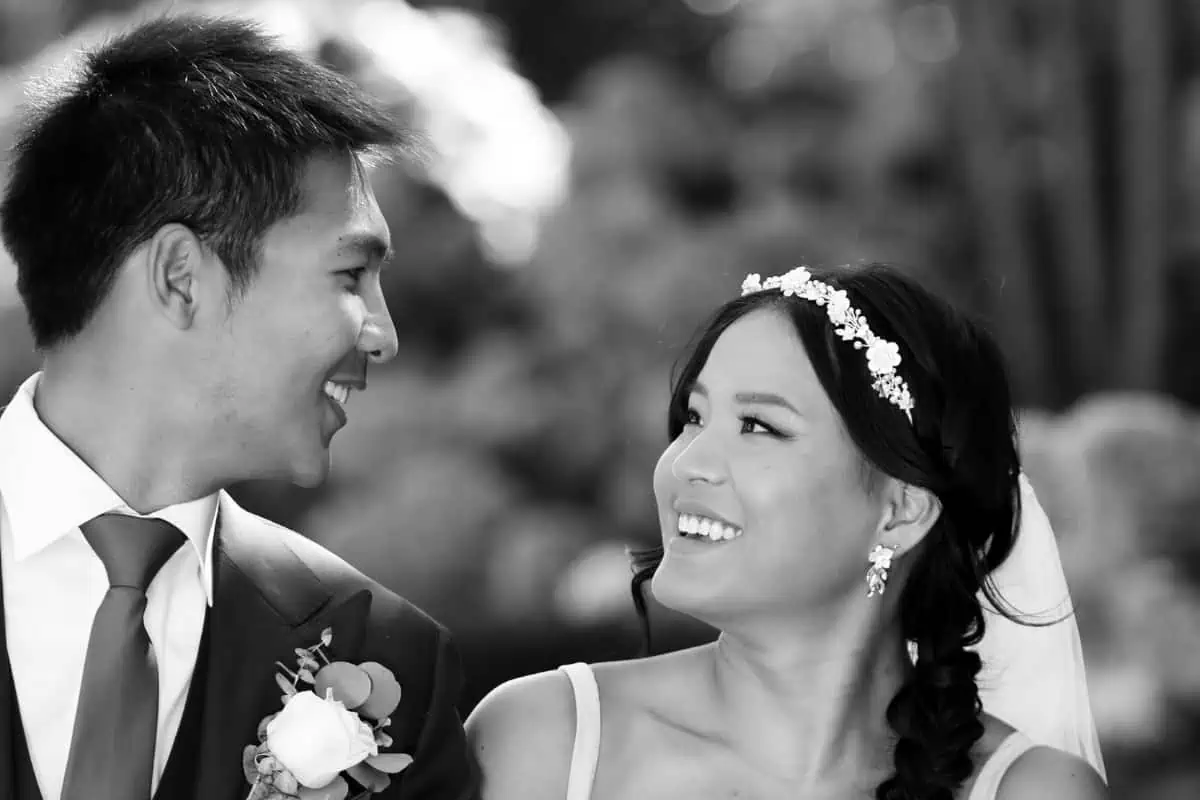 A black and white photo captures a smiling bride and groom, deeply gazing at each other. The groom dons a suit with a boutonniere, while the bride wears a floral headpiece and earrings, her hair elegantly braided. They reminisce about finding perfect wedding venues on Long Island, NY.