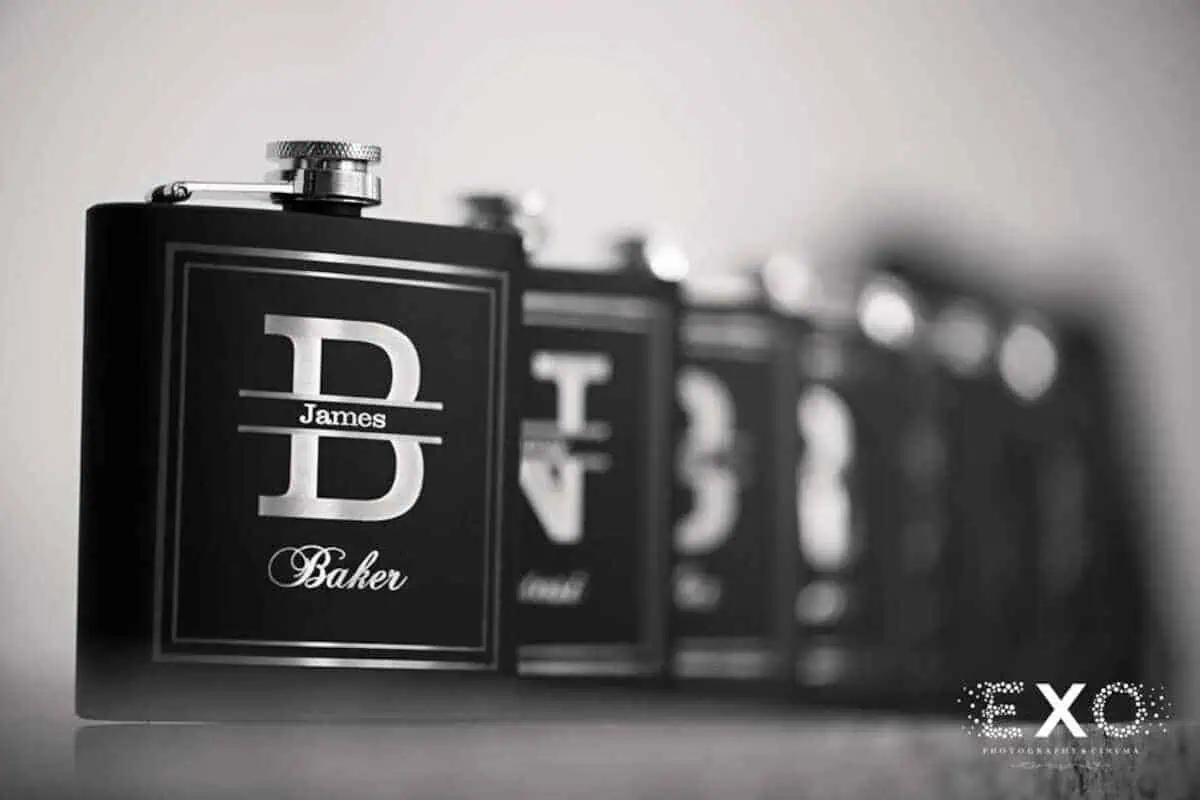 Close-up of a row of black metal flasks with silver accents. The front flask, perfect for groomsmen gifts at wedding venues in Long Island, NY, is labeled with a large "B" and the names "James" and "Baker" in smaller text. The flasks are lined up in a depth-of-field focus.