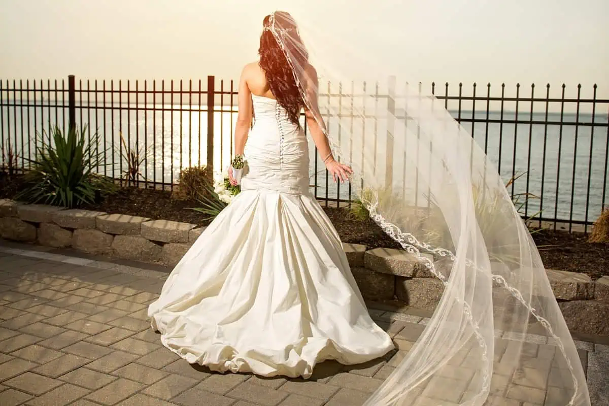 Bridal image of a woman in a white wedding dress, facing away, standing on a brick path by a lake at one of the enchanting wedding venues on Long Island, NY. A long veil flows in the wind as she holds a bouquet. A metal fence and lush plants frame the serene backdrop.