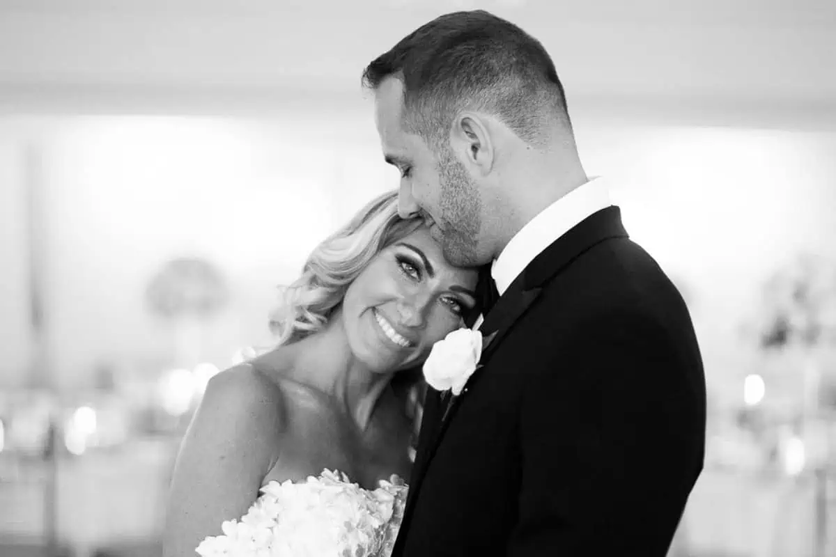 A bride and groom share a happy moment at one of the charming wedding venues on Long Island, NY. The bride, smiling, rests her head on the groom's chest. The groom, in a suit with a boutonniere, leans his head gently against hers. The background is softly blurred in this black and white image.