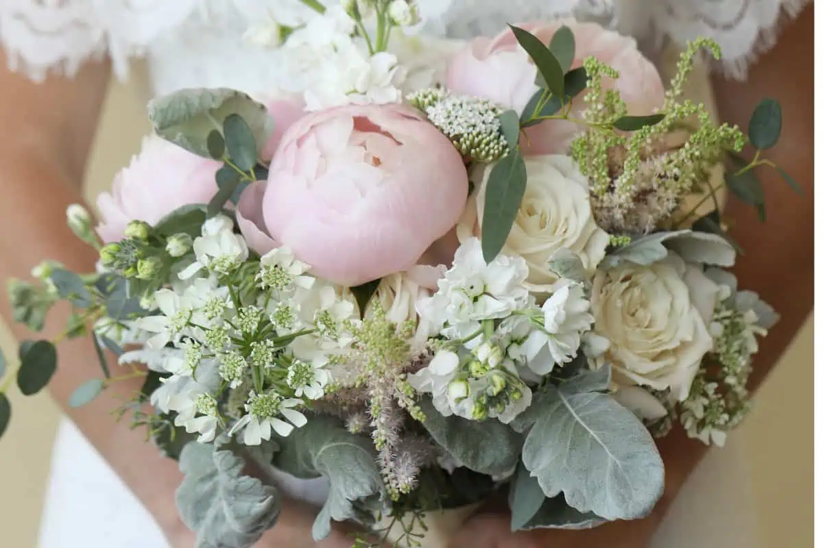 A close-up of a bridal bouquet with large pink peonies, white roses, and greenery like dusty miller and delicate white filler flowers is held in front of a lace bridal gown at one of the charming wedding venues on Long Island, NY.
