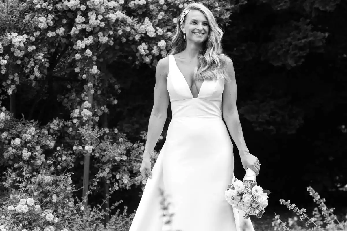 In a classic black and white image, a bride in a flowing white gown stands outdoors at one of the enchanting wedding venues on Long Island, NY. With long, wavy hair and a radiant smile, she is surrounded by blooming flowers and lush greenery, capturing a timeless moment.