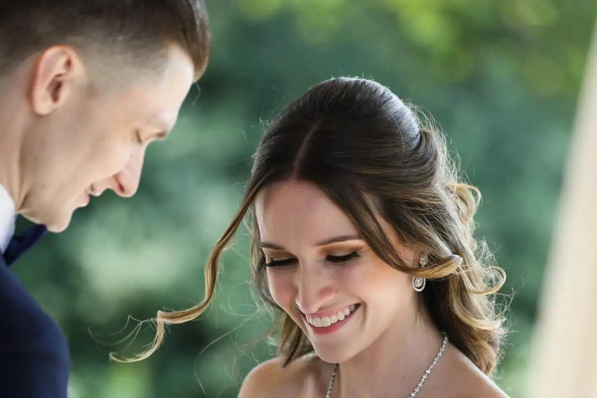 The bride and groom share a joyful moment outdoors at one of the charming wedding venues on Long Island, NY. The bride smiles, looking downwards, with loose curls framing her face. The groom, facing her, also appears joyful. The background is blurred with greenery.