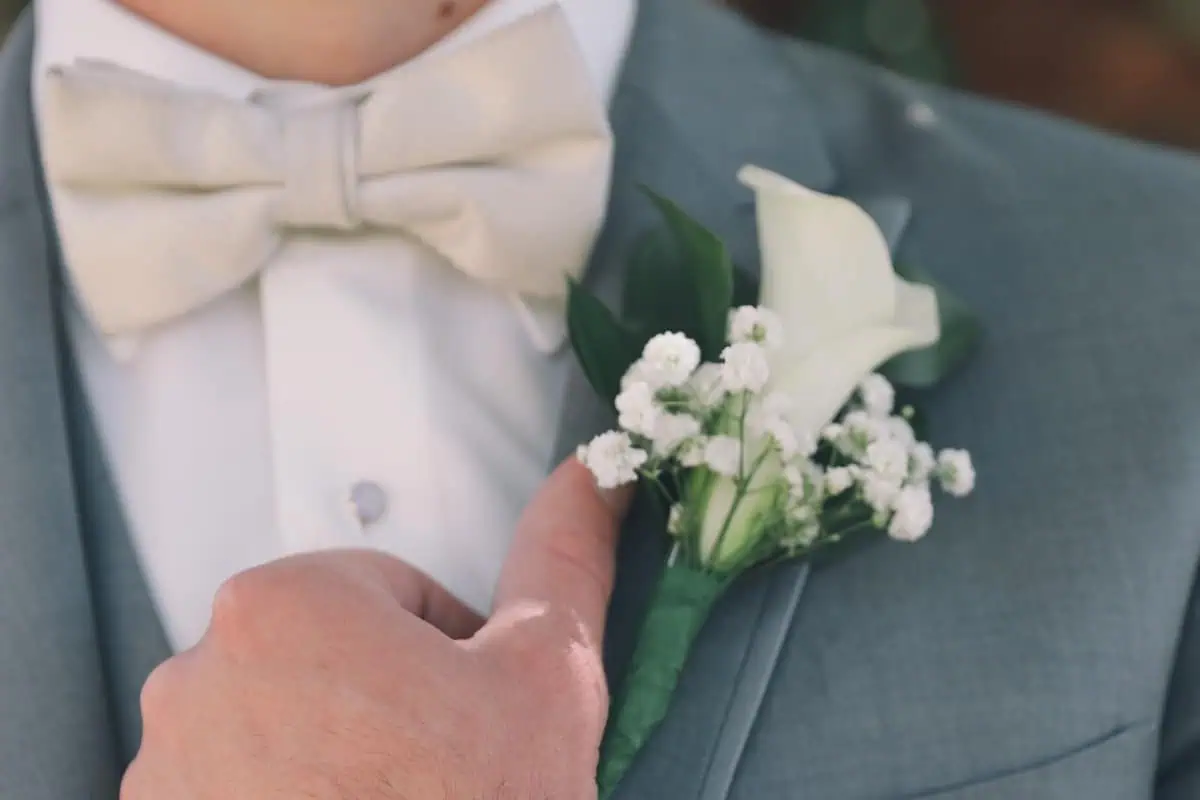 Close-up of a person in a gray suit, white shirt, and cream bow tie adjusting a boutonniere with a white calla lily. This elegant touch is perfect for sophisticated wedding venues on Long Island, NY.