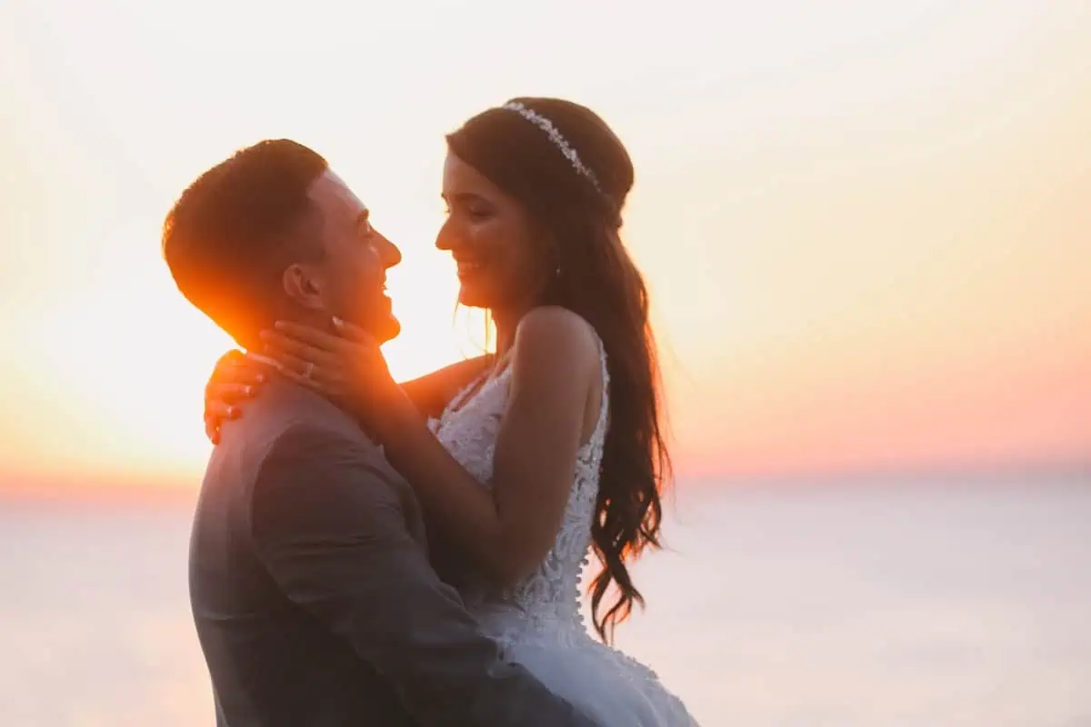 At sunset by the water, a bride and groom embrace joyfully, her white gown and headpiece glowing against the vibrant orange and pink sky. Perfect for capturing memories in one of the enchanting wedding venues on Long Island, NY.