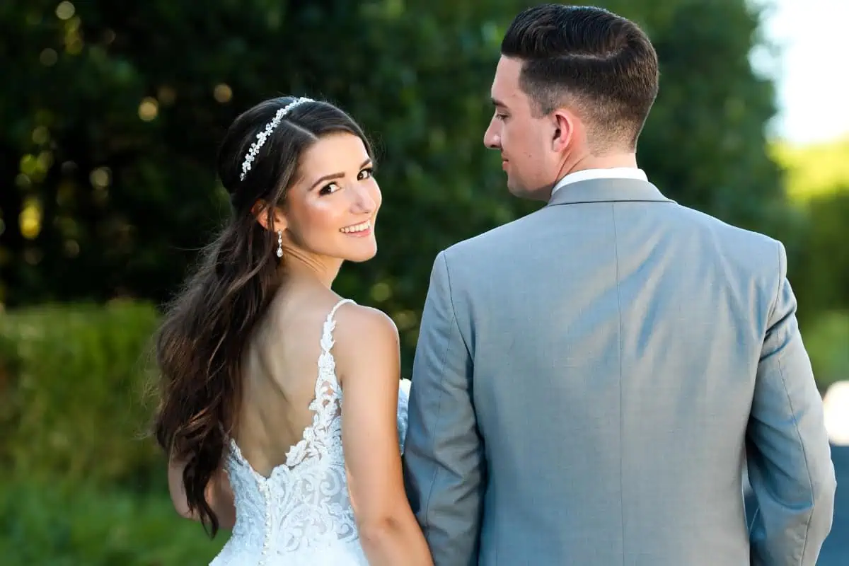 A bride in a white lace gown and a groom in a gray suit stand outdoors, holding hands at one of the stunning wedding venues on Long Island, NY. The bride smiles over her shoulder as the groom gazes adoringly at her, with lush greenery softly blurred in the background.