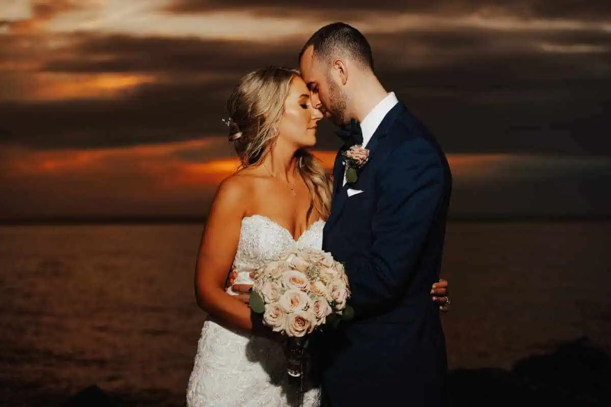 A bride and groom embrace on a beach at sunset, capturing the romance typical of wedding venues in Long Island, NY. The bride’s white lace gown contrasts beautifully with her bouquet of roses, while the groom's dark suit stands out against a sky filled with dramatic clouds and orange hues.