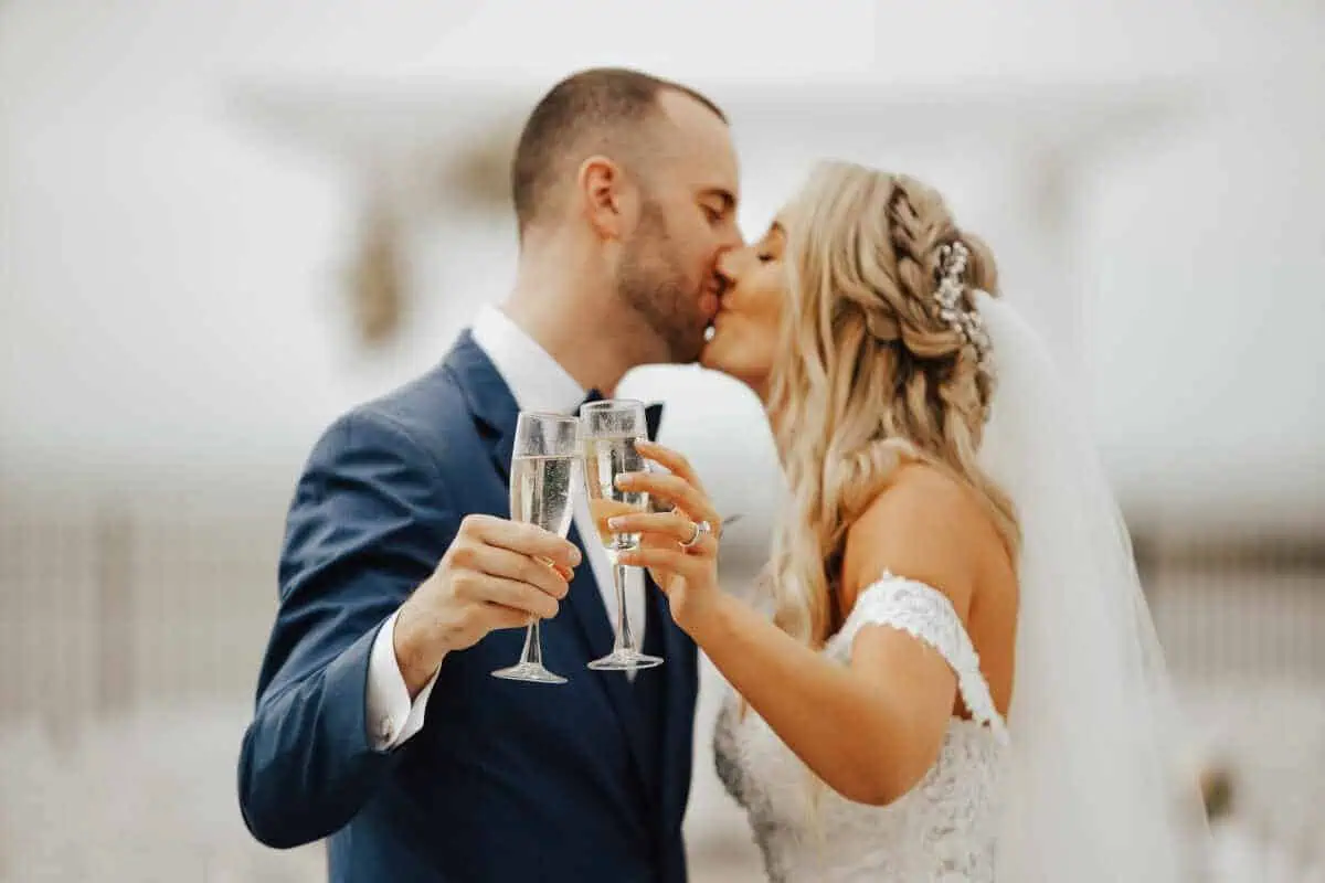 A bride and groom kiss while holding champagne glasses, celebrating their love at one of the charming wedding venues on Long Island, NY. The groom is in a blue suit, and the bride wears an off-the-shoulder white lace dress with her hair elegantly adorned. They appear to be outdoors.
