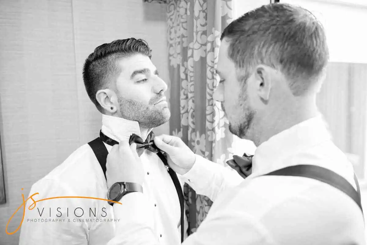 Two men in formal attire, one adjusting the other's bow tie, are likely preparing for a special occasion. Indoors with patterned curtains in the background, they focus intently on perfecting the bow tie arrangement—perhaps a detail for an event at one of the exquisite wedding venues Long Island NY offers.