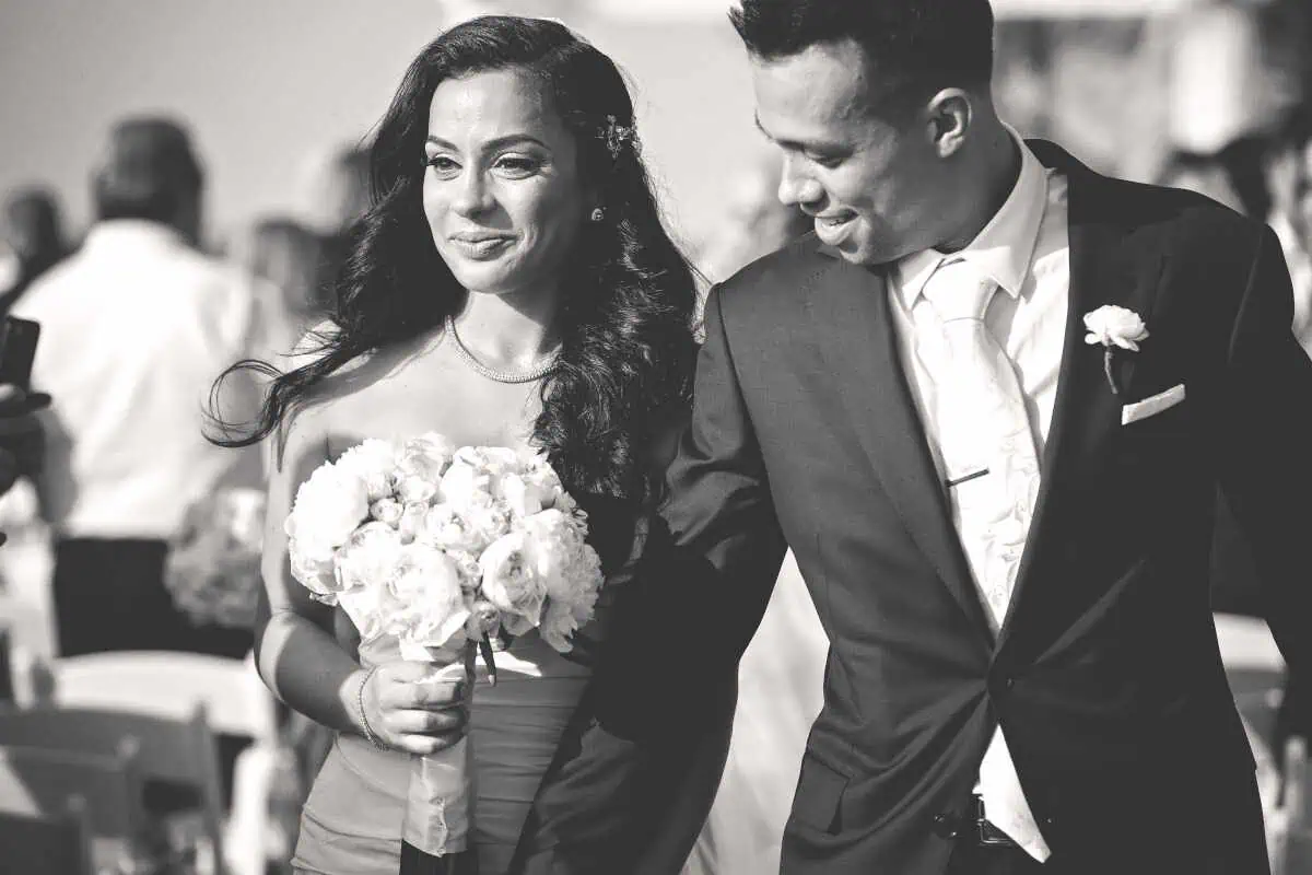 A black and white photo captures a joyful couple at a wedding on Long Island, NY. The woman holds a bouquet, both are beaming in formal attire, surrounded by standing guests at one of the charming wedding venues in the area.