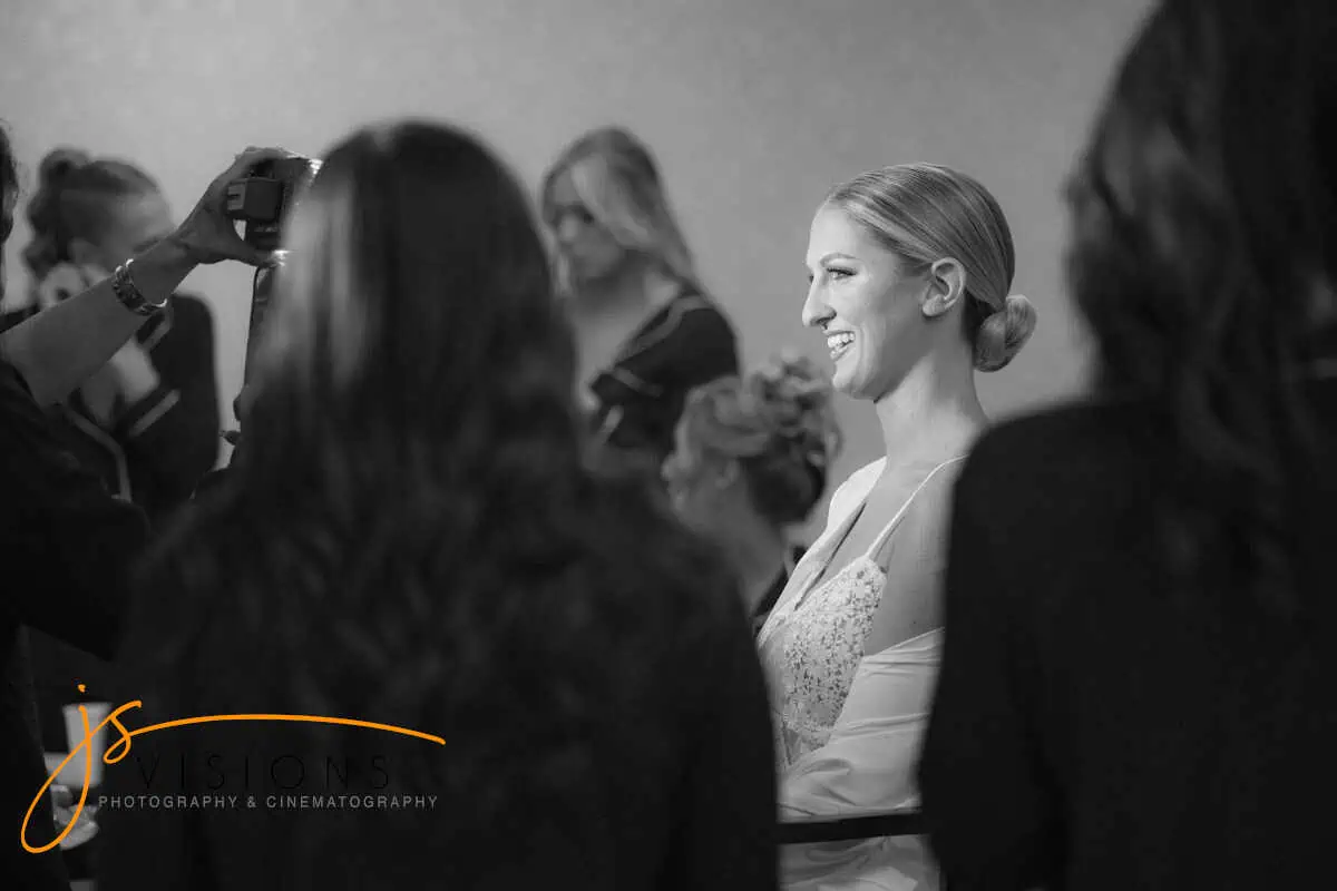A bride with a bright smile, seated in a sparkling gown, is surrounded by her group at one of the charming wedding venues Long Island NY offers. She faces someone taking her photo as others prepare in the background, creating a joyful atmosphere. Black and white photography with a logo at the bottom.