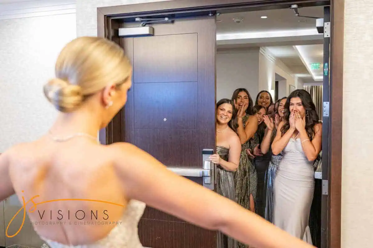 A bride, seen from behind in a white gown, faces a doorway at one of the stunning wedding venues on Long Island, NY. There, a group of excited women dressed in formal attire expresses delight and surprise.