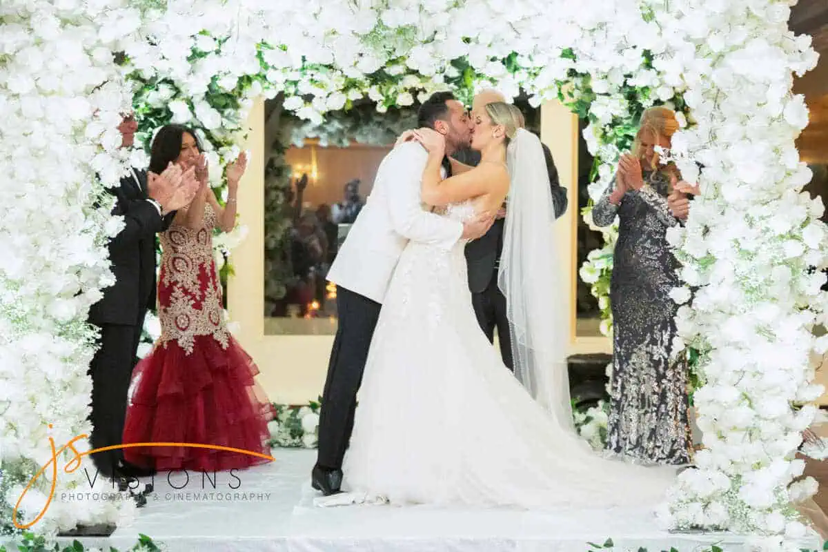 A bride and groom share a kiss under a floral arch at their wedding ceremony, held at one of the picturesque wedding venues on Long Island, NY. The bride wears a flowing white gown and veil, while the groom sports a white tuxedo jacket. Guests around them are clapping and smiling, enhancing the bright, joyful scene.