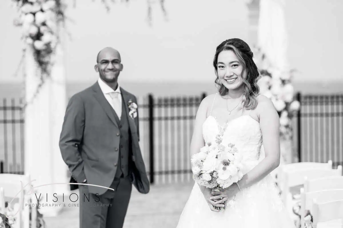 A bride in a white gown holds a bouquet, smiling at the camera, while a groom in a suit stands behind her with equal joy. Captured outdoors at one of the picturesque wedding venues on Long Island, NY, they stand near a decorative arch and chairs. The image is beautifully in black and white.