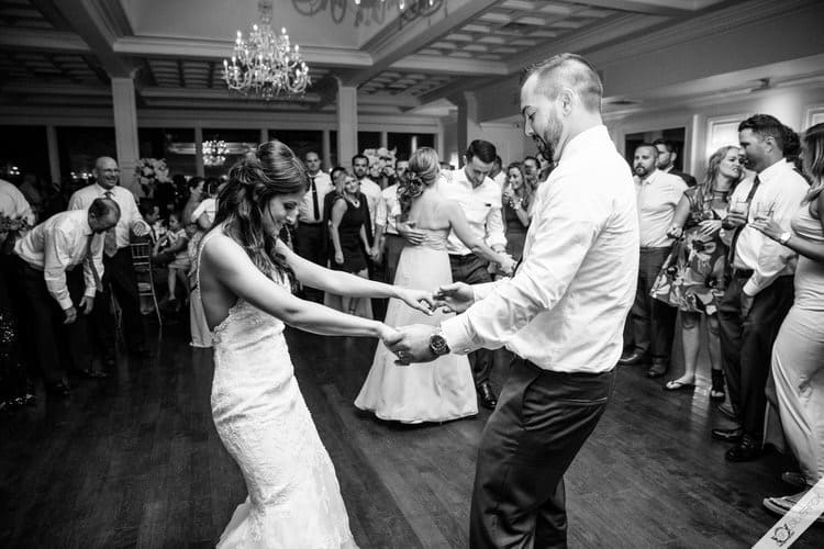A bride and groom dance joyfully in the center of a crowded ballroom, a classic choice among wedding venues on Long Island, NY. The bride wears a white gown and the groom is in a white shirt. Guests watch, smiling and dressed in formal attire beneath elegant chandeliers. Black and white photo.