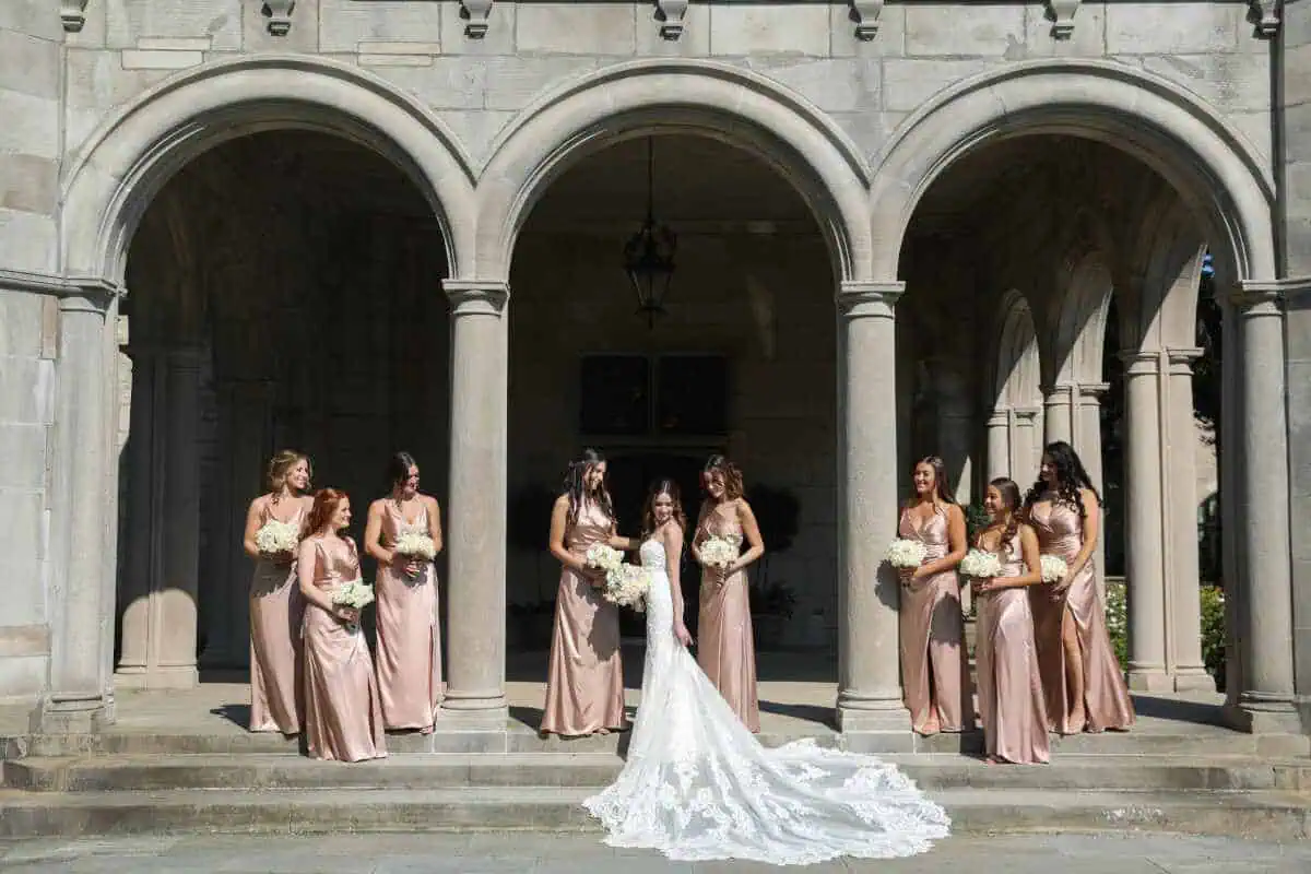 A bride stands with eight bridesmaids under an archway at one of the elegant wedding venues in Long Island, NY. She wears a white gown with a long train, holding a bouquet. The bridesmaids wear matching satin dresses, carrying white bouquets against the backdrop of the stone building's arches.