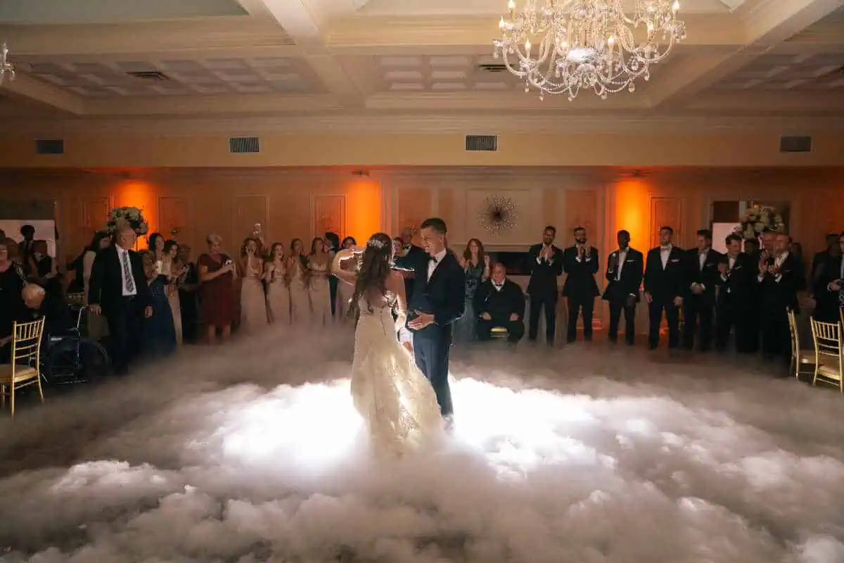 A bride and groom share their first dance on a cloud-like floor with fog, surrounded by guests in formal attire. A chandelier hangs from the ceiling, illuminating the elegant ballroom with warm lighting—a perfect scene often found in wedding venues on Long Island, NY.