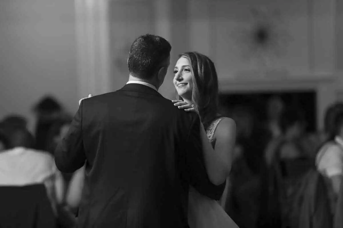 In a dimly lit room at one of the enchanting wedding venues on Long Island, NY, a couple dances closely. The woman smiles at her partner, surrounded by softly blurred figures that enhance the romantic atmosphere. The timeless scene is captured in black and white.