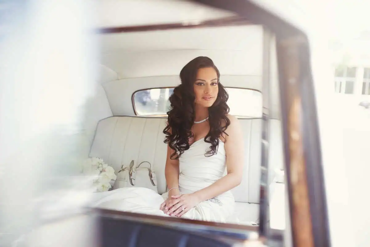 A bride with long, dark, wavy hair sits in the back of a vintage car, heading to one of the charming wedding venues on Long Island, NY. She is wearing a strapless white wedding dress and a necklace, smiling softly, with a bouquet of white flowers beside her. The car's interior is visible.