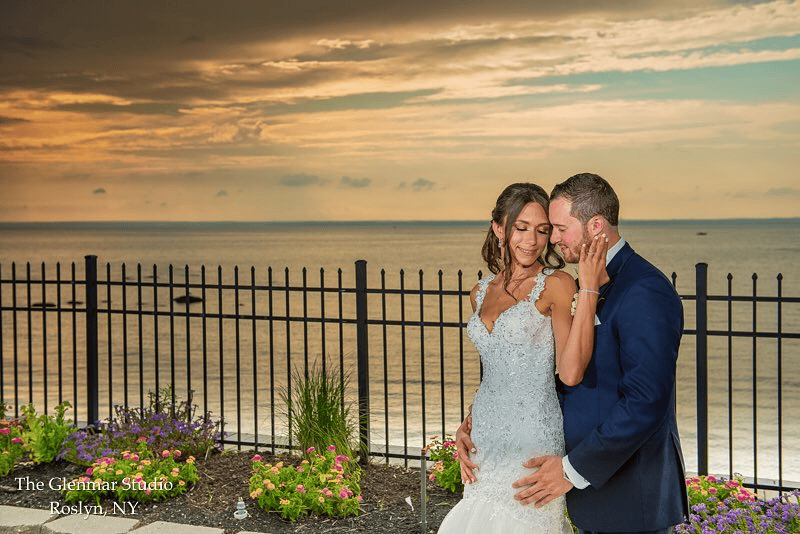A smiling bride and groom embrace near a fence with a scenic ocean backdrop, celebrating their anniversary. The sky is cloudy, with a warm sunset in the distance. The garden in front features colorful flowers. Text at the bottom left reads "The Glentar Studio, Roslyn, NY.