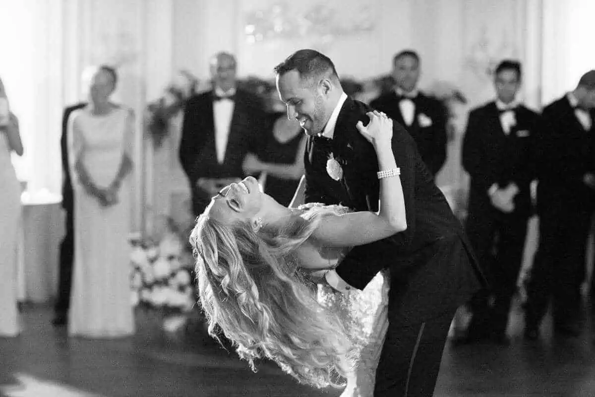 A bride and groom share a joyful dance, with the groom gently dipping the bride as they both smile. Guests in formal attire watch in the background, enveloped by an elegantly decorated ambiance typical of wedding venues on Long Island, NY.