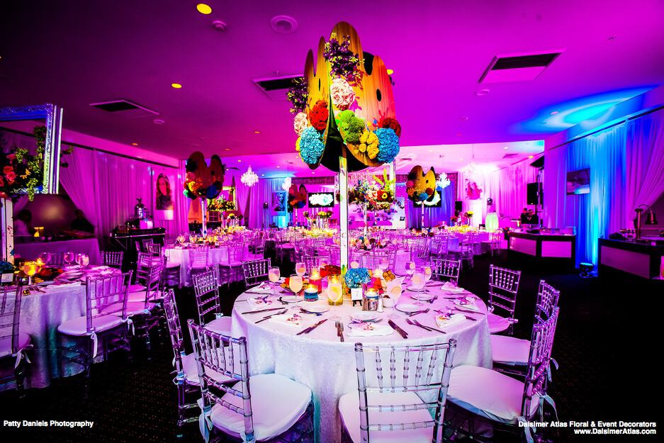 A vibrant banquet hall, ideal for wedding venues on Long Island, NY, features round tables adorned with white cloths and clear chairs. Colorful floral centerpieces glow under pink and purple lighting, enhancing the festive atmosphere. Plates and glasses are neatly arranged on tables.