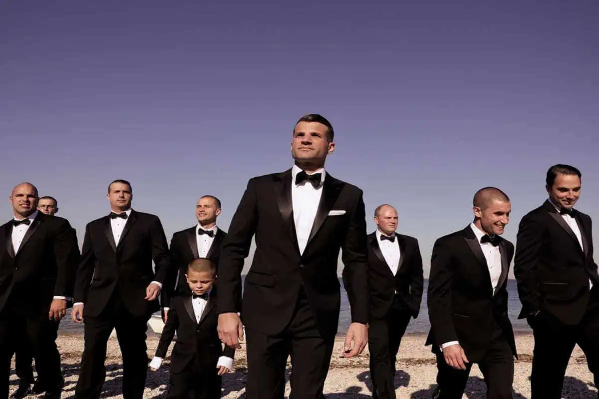 A group of men and a young boy, all in black tuxedos with bow ties, walk confidently on a beach under a clear blue sky. The man in the center stands slightly ahead, exuding leadership—an elegant scene reminiscent of wedding venues on Long Island NY.
