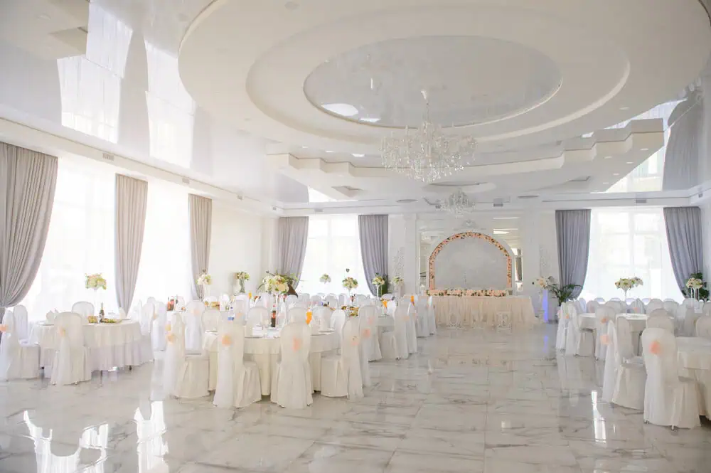 A spacious and elegantly decorated banquet hall in Long Island, NY, perfect for wedding venues. White tablecloth-covered round tables with chairs, soft lighting, tall draped windows, a chandelier, and floral arrangements create a luxurious ambiance.