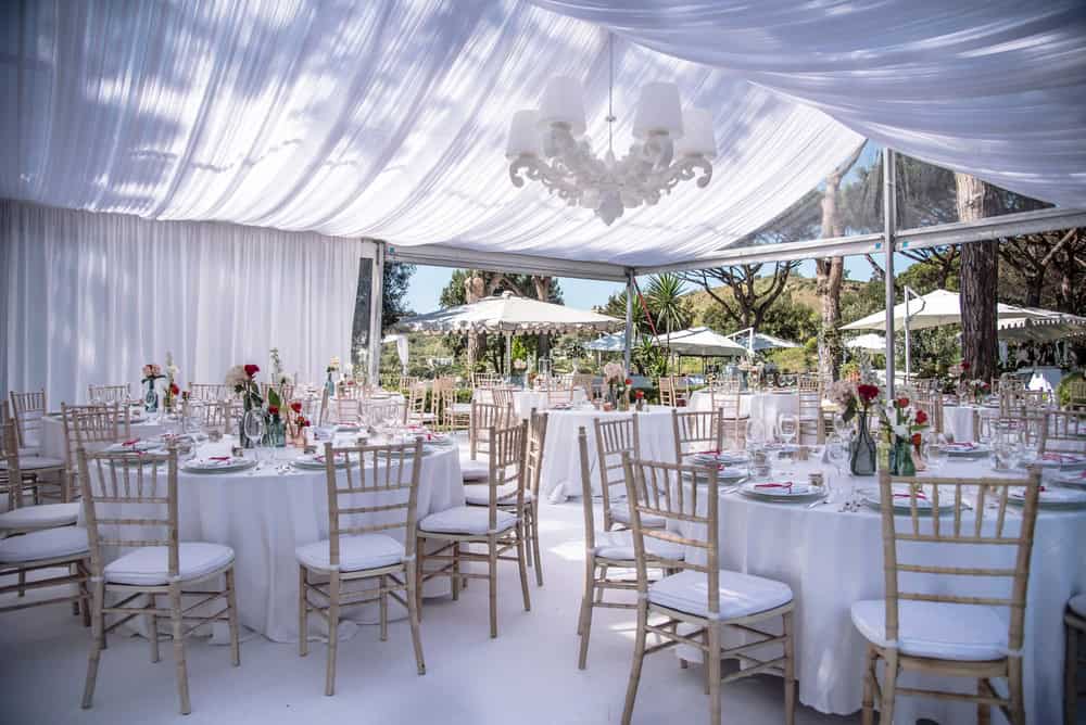 Elegant outdoor wedding reception setup under a white draped canopy with round tables, white tablecloths, and wooden chairs. Tables are decorated with red and white floral arrangements. Wedding venues on Long Island, NY often feature large windows showcasing a garden view.