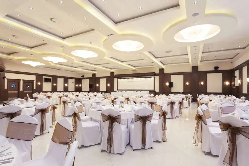A spacious ballroom, ideal for wedding venues on Long Island, NY, is set for an event. It features round tables with white tablecloths and chairs adorned with elegant covers and brown bows. Modern lighting fixtures grace the ceiling, and a large screen is visible at one end of the room.