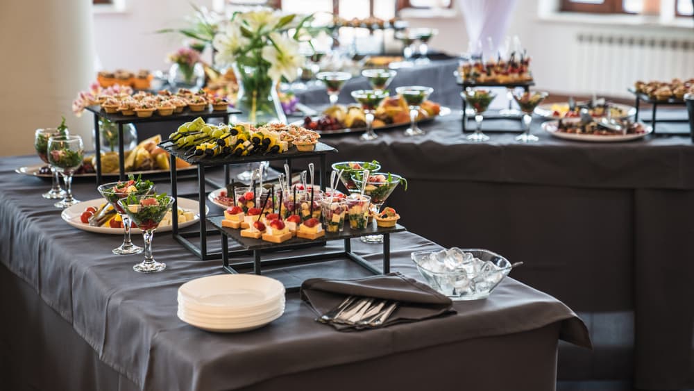 A buffet table adorned with a variety of appetizers—skewers, small sandwiches, and glass-served salads—graces the wedding venues in Long Island, NY. Set on black-clothed tables with neatly arranged plates and cutlery, it’s complemented by an elegant floral centerpiece.