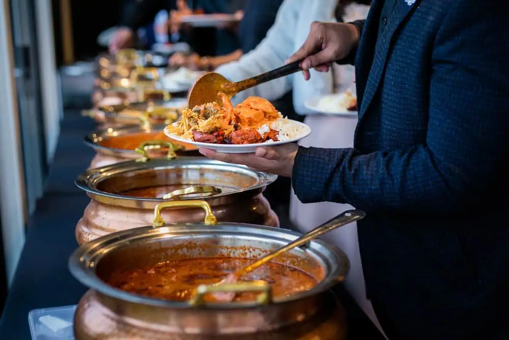 A person in a dark jacket serves themselves from a buffet of Indian dishes in large copper pots, perhaps at one of the elegant wedding venues on Long Island, NY. Their plate is filled with a variety of food items, while several others help themselves in the background.