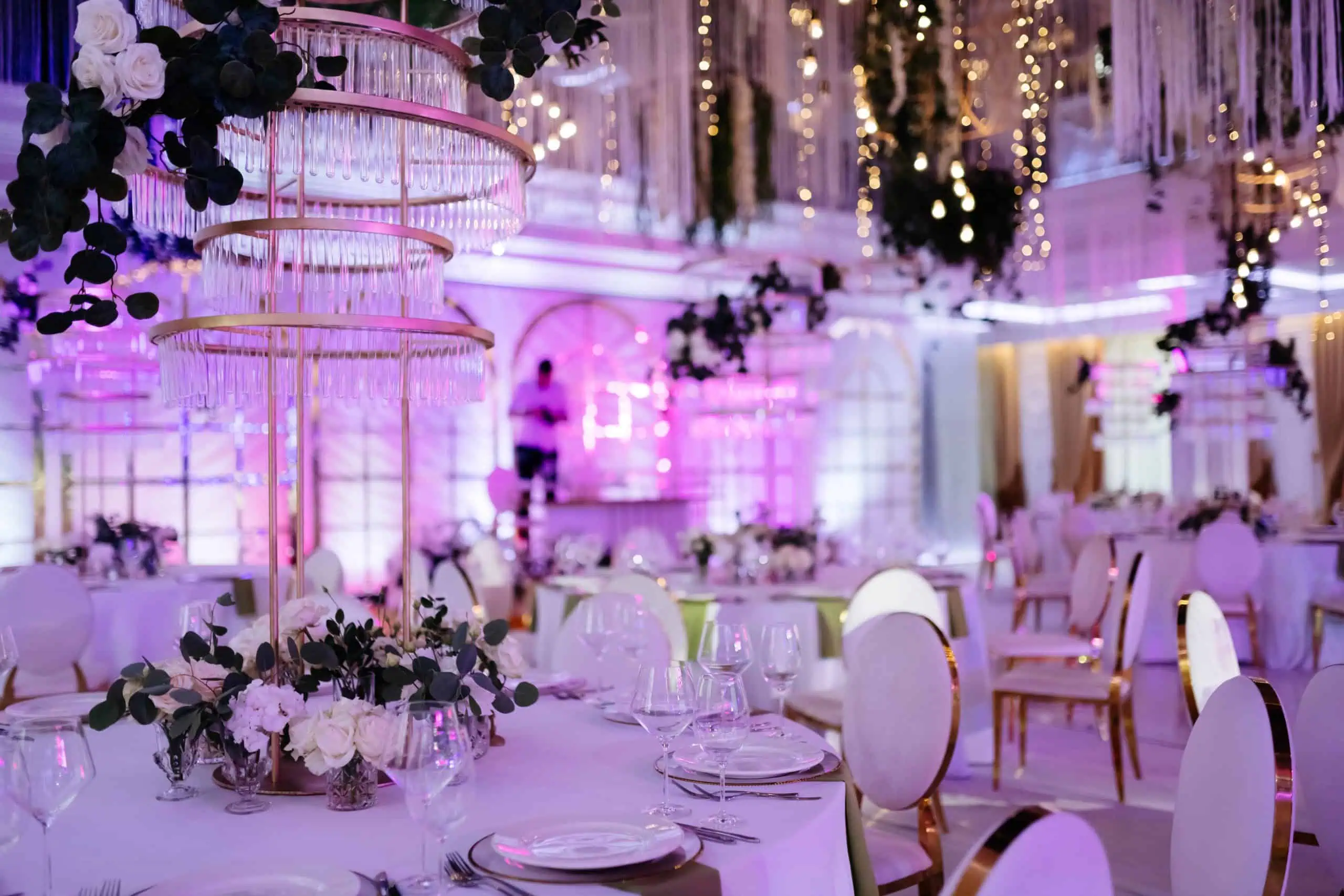 Elegant wedding reception setup at one of the finest wedding venues in Long Island, NY, featuring round tables, white chairs, floral centerpieces, and crystal chandeliers. The room is lit with pink and purple lighting, draped with hanging decorations and fairy lights for a romantic ambiance.