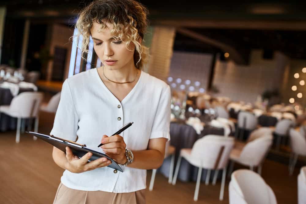 A person with curly hair stands in an elegantly set dining area, holding a clipboard and writing. They are wearing a white shirt and appear focused, likely managing or organizing one of the charming wedding venues Long Island NY has to offer.