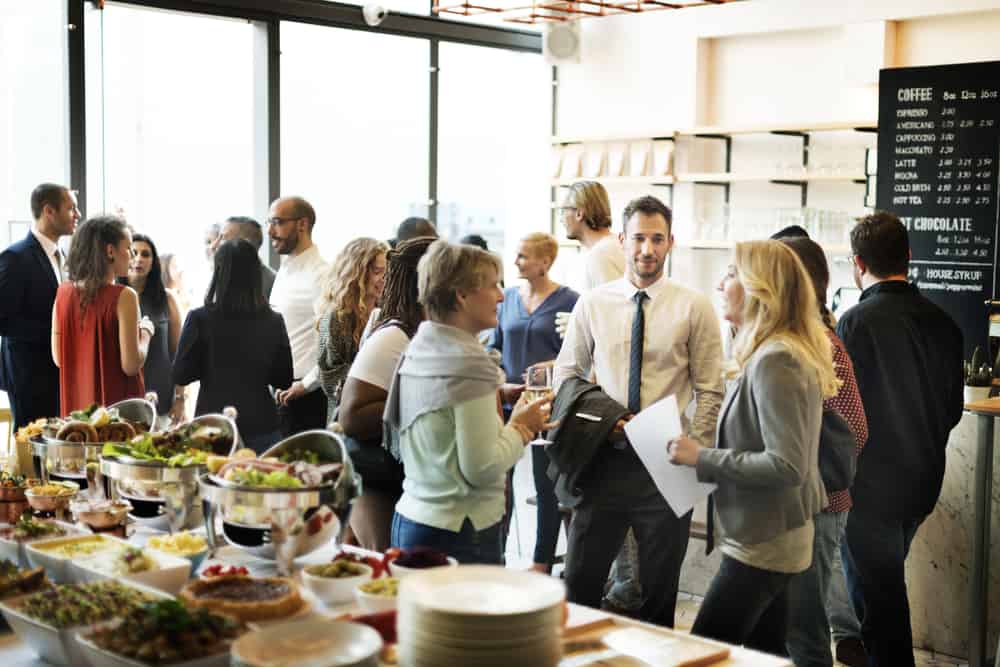 A group of people socializing in a cafe-like setting, reminiscent of cozy wedding venues on Long Island, NY. Some gather near a table with bowls of salad and plates of food. A large menu board lists beverages. The atmosphere is lively and casual, with natural light streaming through large windows.