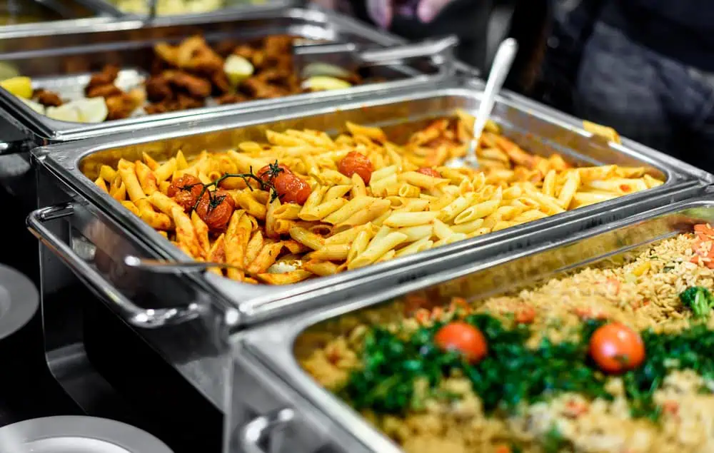 Buffet with trays of food features penne pasta with tomatoes, a rice dish with greens and tomatoes, and a dish with breaded items, perfect for weddings. A serving spoon rests in the pasta tray, ready to serve guests at wedding venues on Long Island, NY.