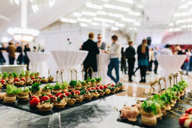 Trays of assorted appetizers with garnishes adorn a table in the foreground; in the background, guests mingle in a spacious, well-lit wedding venue on Long Island, NY, featuring high ceilings and elegant round standing tables.