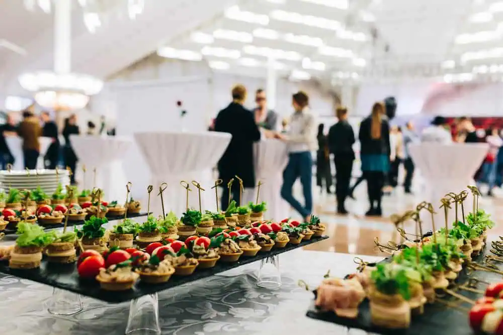 Trays of assorted appetizers with garnishes adorn a table in the foreground; in the background, guests mingle in a spacious, well-lit wedding venue on Long Island, NY, featuring high ceilings and elegant round standing tables.