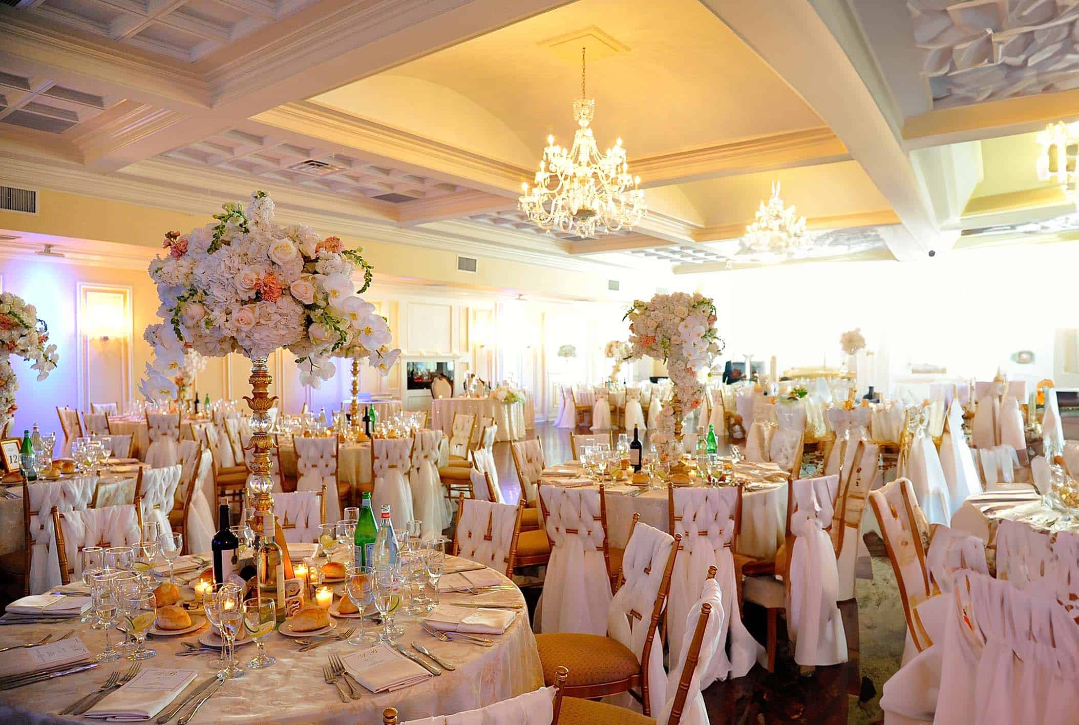 An elegant banquet hall on Long Island, NY, is set for a wedding reception. Tables are adorned with white linens, floral centerpieces of various blooms, and candles. Chandeliers hang from the ceiling, casting soft light over this luxurious and formal wedding venue.