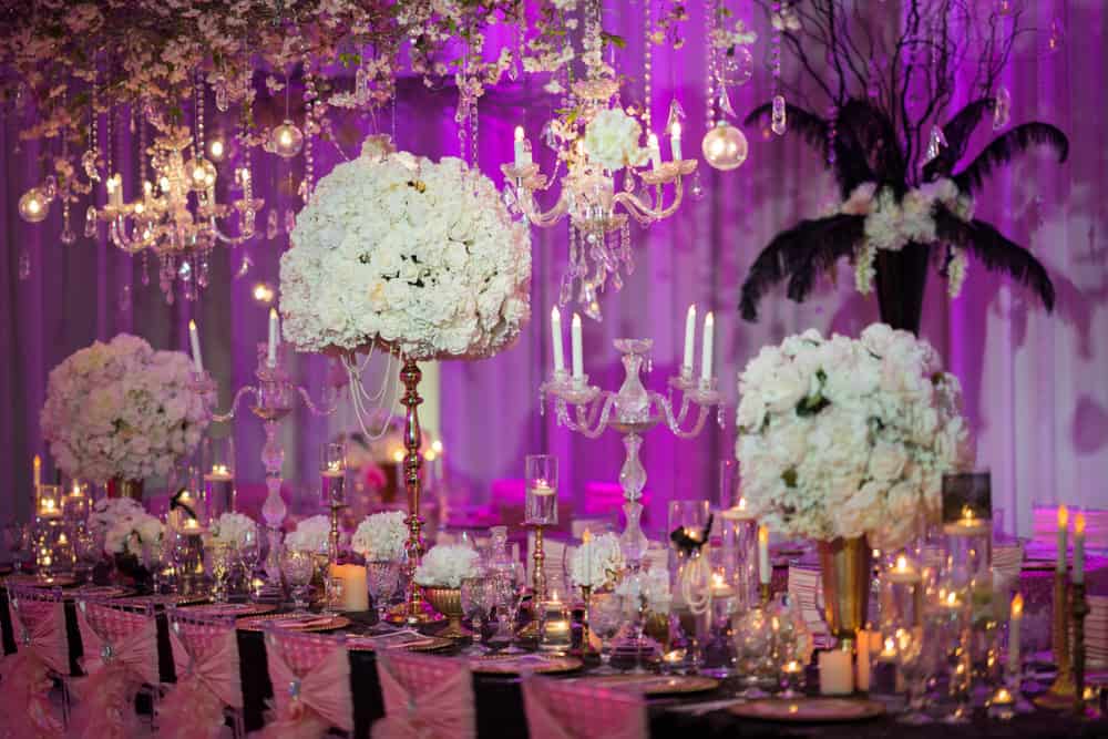 Elegant wedding reception setup with large floral centerpieces, crystal chandeliers, and candles on a long table. Pink and purple lighting creates a romantic ambiance. Draped fabric and glassware add to the luxurious decor—perfect for those seeking wedding venues Long Island NY offers.