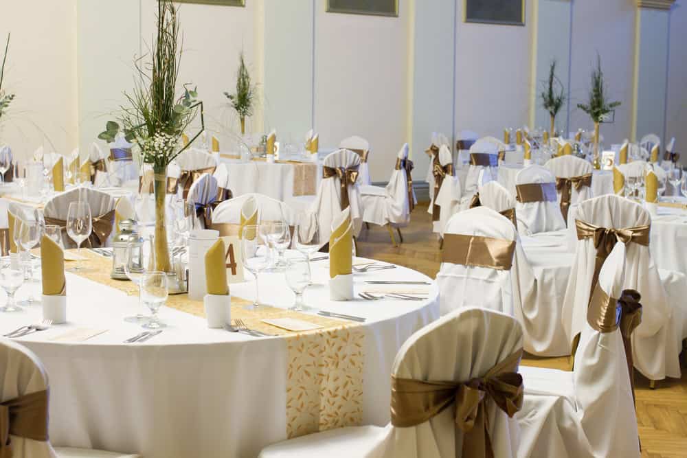 A beautiful wedding reception setup in one of the elegant wedding venues on Long Island, NY. Round tables draped with white tablecloths and brown sashes on chairs host tall centerpieces with greenery, complemented by glasses, napkins, and silverware for a perfect celebration.