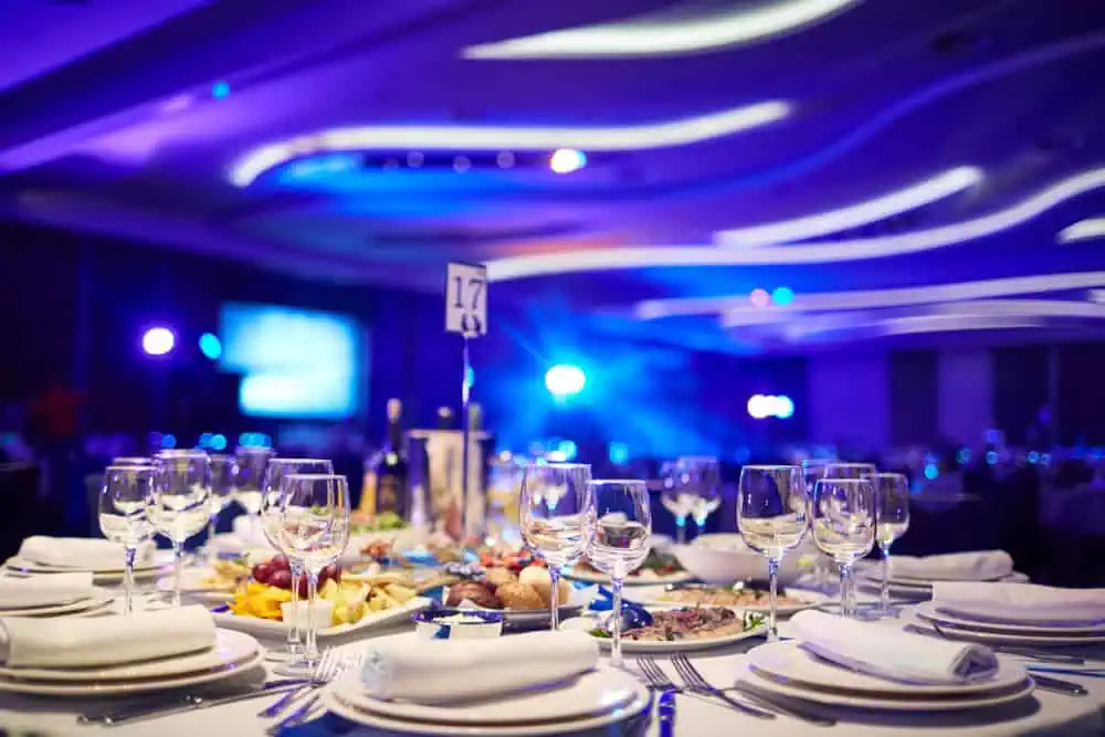A lavishly set banquet table with elegant dinnerware and glasses, featuring plates of food. The room, reminiscent of top wedding venues on Long Island, NY, is dimly lit with blue ambient lighting and a blurred background hinting at a stage, creating a sophisticated event atmosphere.