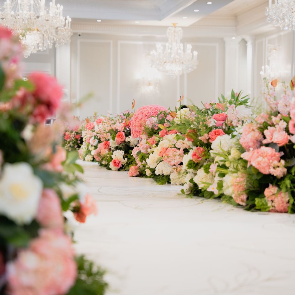 A bright, elegant ballroom decorated with luxurious chandeliers and lush arrangements of pink, peach, and white flowers lining a central aisle.