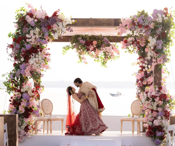 A couple in traditional Indian wedding attire shares a dip pose under a floral archway, with chairs on each side and a serene waterfront background.